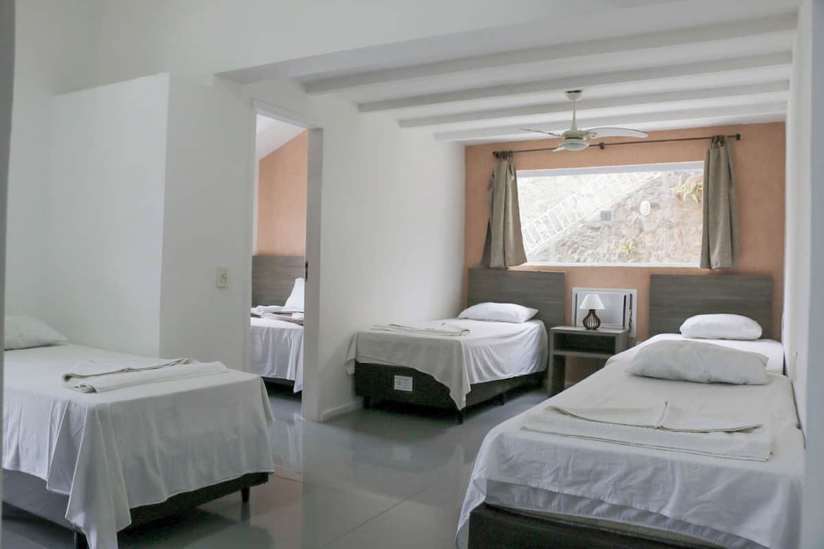 A hotel room with three single beds, a window with curtains, a ceiling fan, and a small table with a lamp.