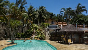 Una piscina con plataforma de piedra, rodeada de palmeras y una estructura con techo de paja.