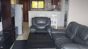 Een woonkamer met een zwarte leren bank, een bijzettafeltje en een tv-meubel. Achtergrond is een keuken met kasten en een raam.