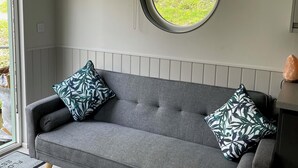 Een grijze bank met twee gekleurde kussens, een raam met een groene zonwering, en een kleine verwarming op een tafel.