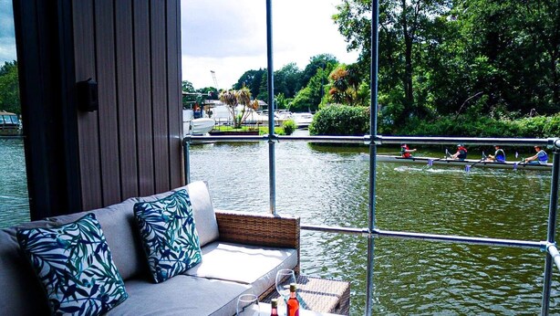 Een moderne buitenruimte met een bank en tafel, met uitzicht op een rustige rivier waar geroeid wordt.