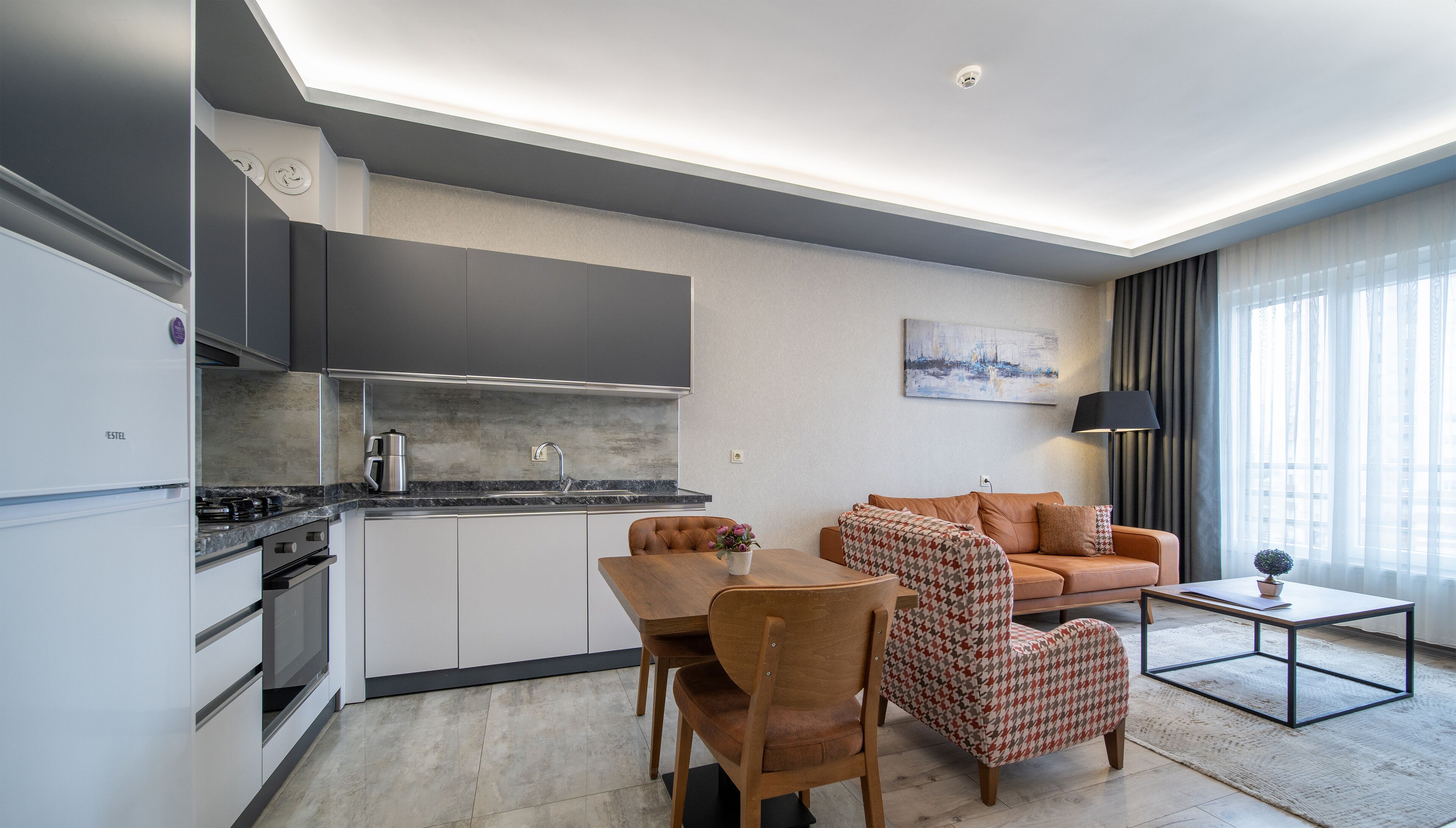 A modern kitchen with a built-in oven, a coffee maker, and a sink. There is a dining area with a table, chairs, and a sofa. A lamp is on a side table, and a painting is on the wall.