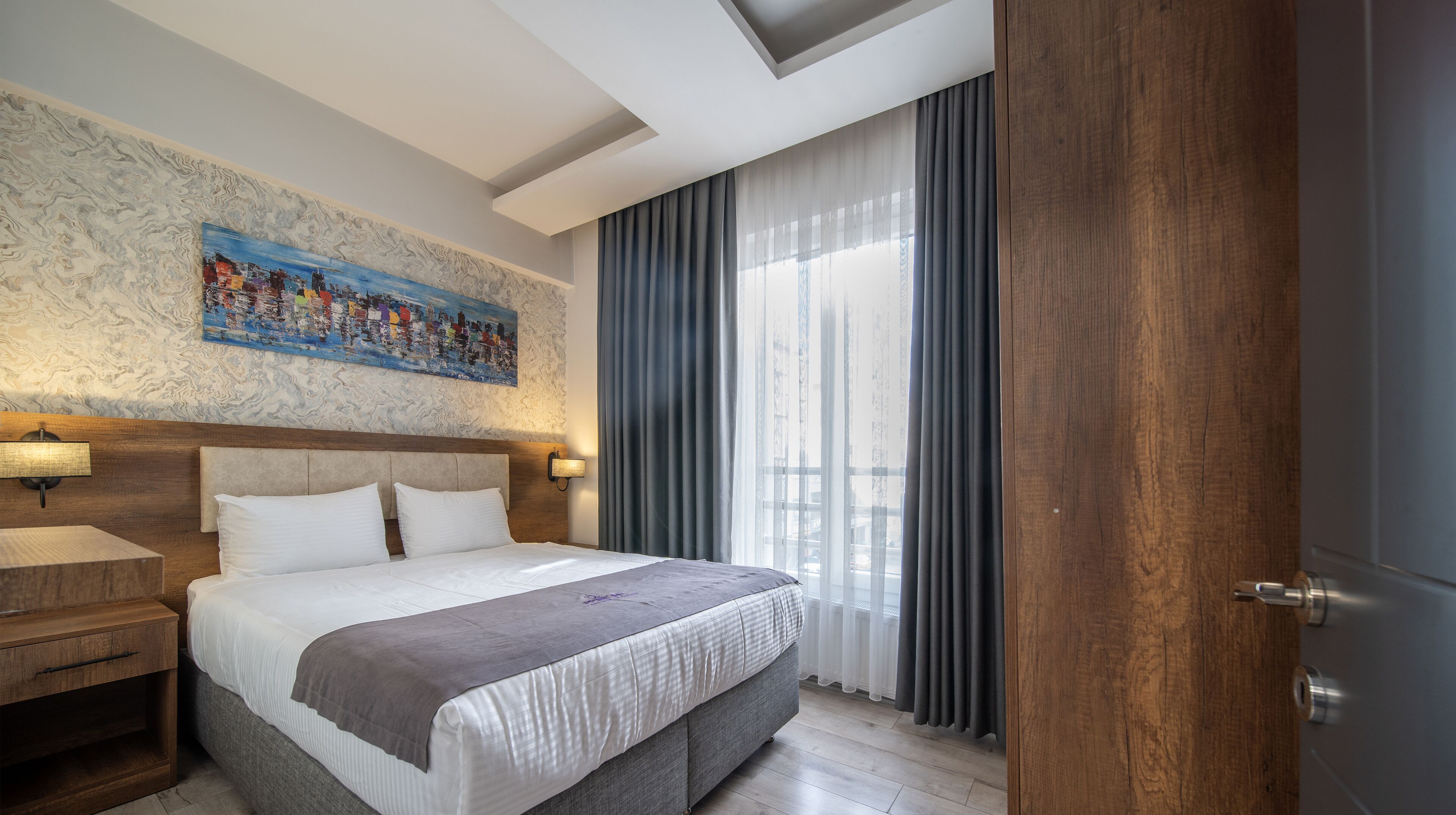 Habitación de hotel con una cama grande, cabecera con una pintura de un paisaje urbano, mesita de noche de madera y vistas al exterior a través de cortinas traslúcidas.