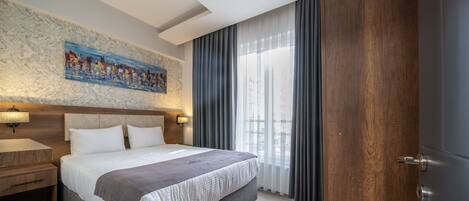 Habitación de hotel con una cama grande, cabecera con una pintura de un paisaje urbano, mesita de noche de madera y vistas al exterior a través de cortinas traslúcidas.