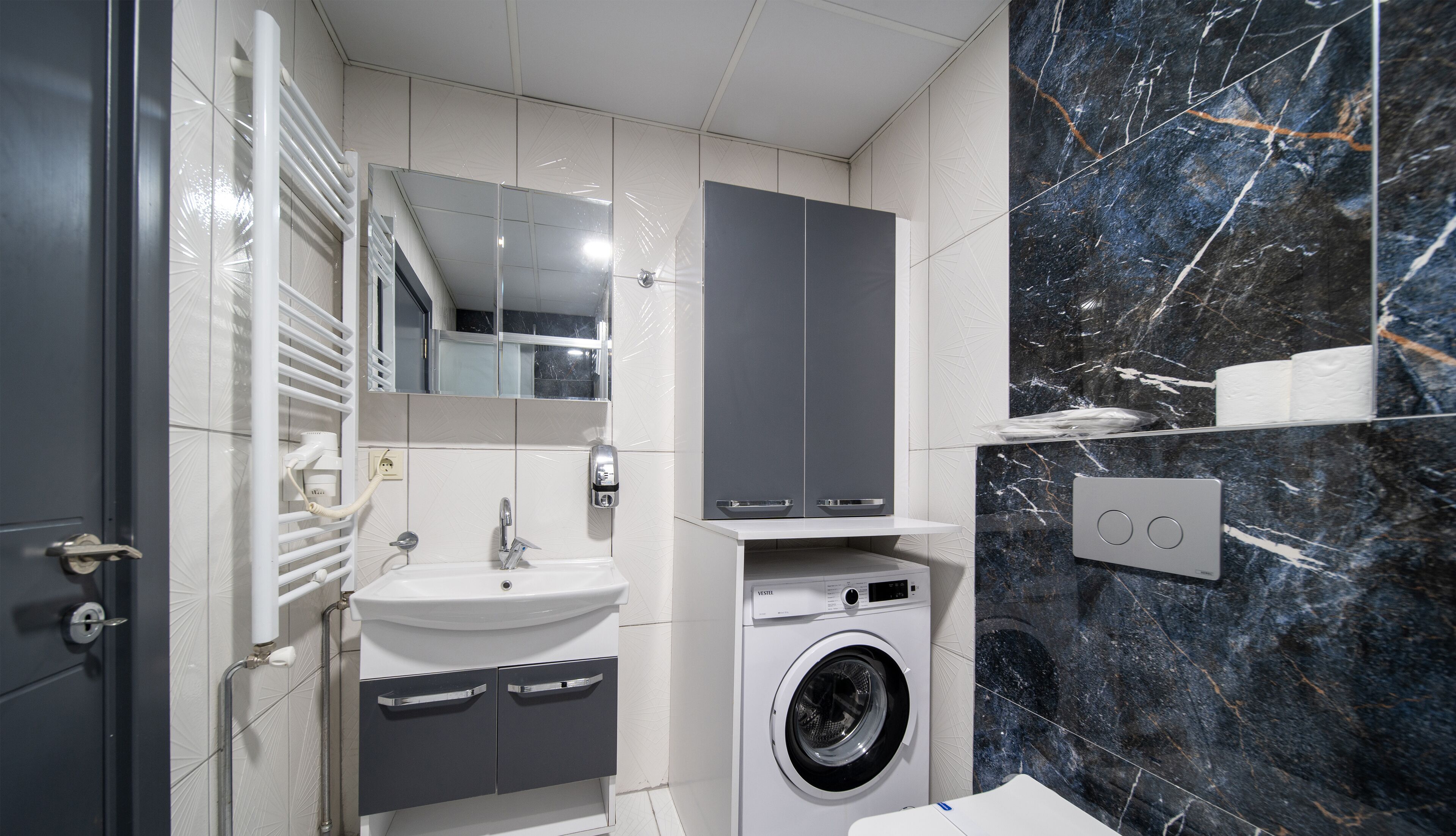 A modern bathroom with a white sink, a built-in washing machine, a mirror, and a marble backsplash.