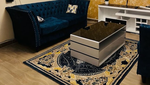 Ein Wohnzimmer mit einem dunkelblauen Sofa, einem Glastisch, einem gemusterten Teppich und einem Fernseher auf einem weißen Schrank.