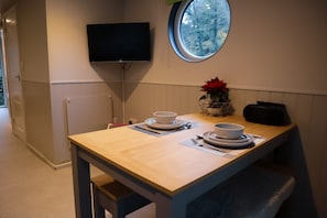 A small dining area with a wooden table set for two, a television mounted on the wall, and a window with a view of trees.