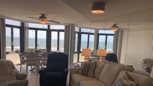 A spacious living room with a large window overlooking the ocean, a ceiling fan, and a cozy seating area with a sofa and armchairs. - 3 bedroom, 3 bath beautiful oceanfront condo on the quiet shore of Cherry Grove. (North Myrtle Beach)