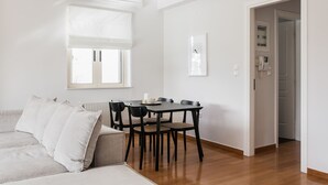 A modern living room with a sofa, a dining table with chairs, a window with blinds, and a door leading to another room. - Exquisite 2BR Apartment (Agia Paraskevi)