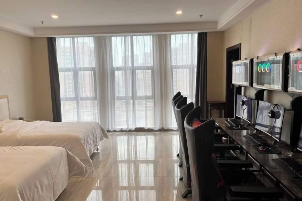 Camera d'albergo con due letti, una scrivania con più monitor e ampie finestre con tende leggere.