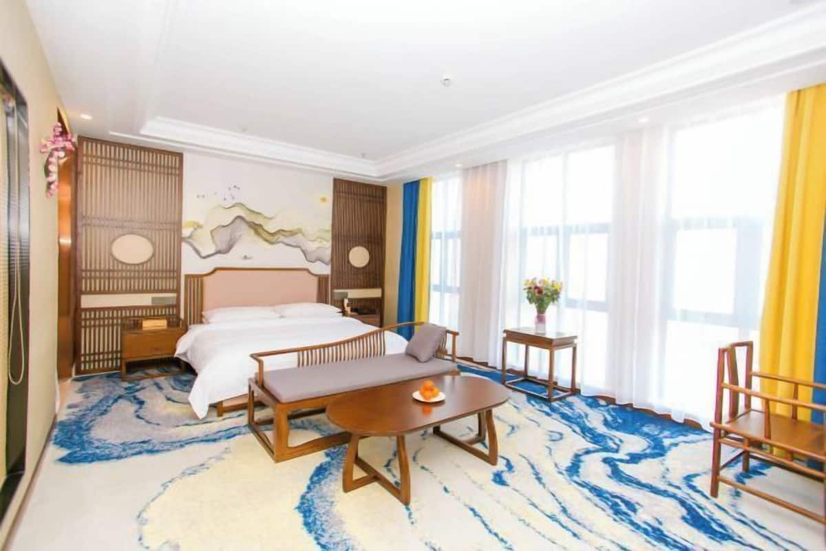 Camera d'albergo con un letto grande, un divano, un tavolino e una sedia. Alle finestre sono presenti tende e un pannello decorativo.