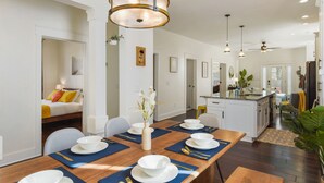Uma sala de jantar com mesa de madeira posta para quatro pessoas, um vaso com flores e uma cozinha ao fundo.