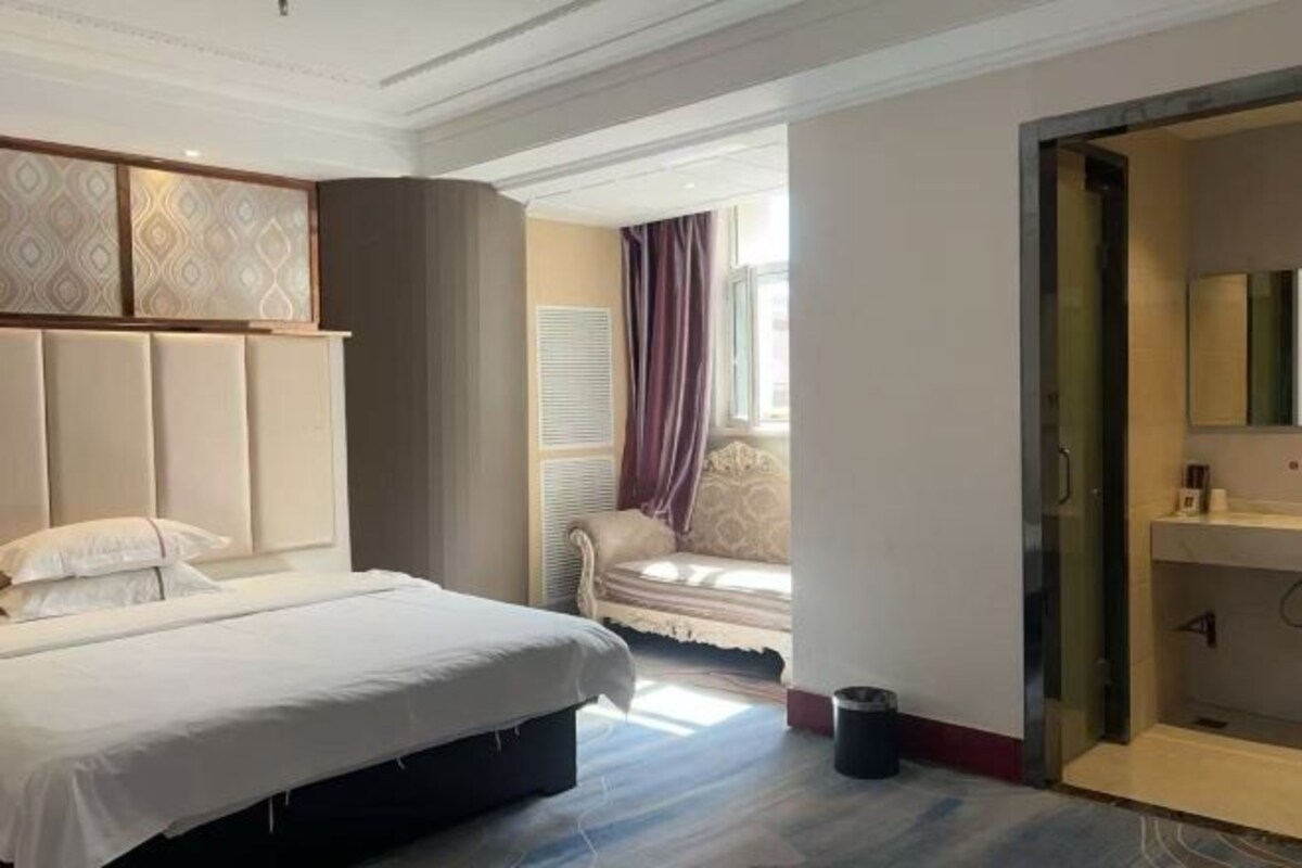 A hotel room with a bed, a chair by the window, a mirror, and a door leading to a bathroom.