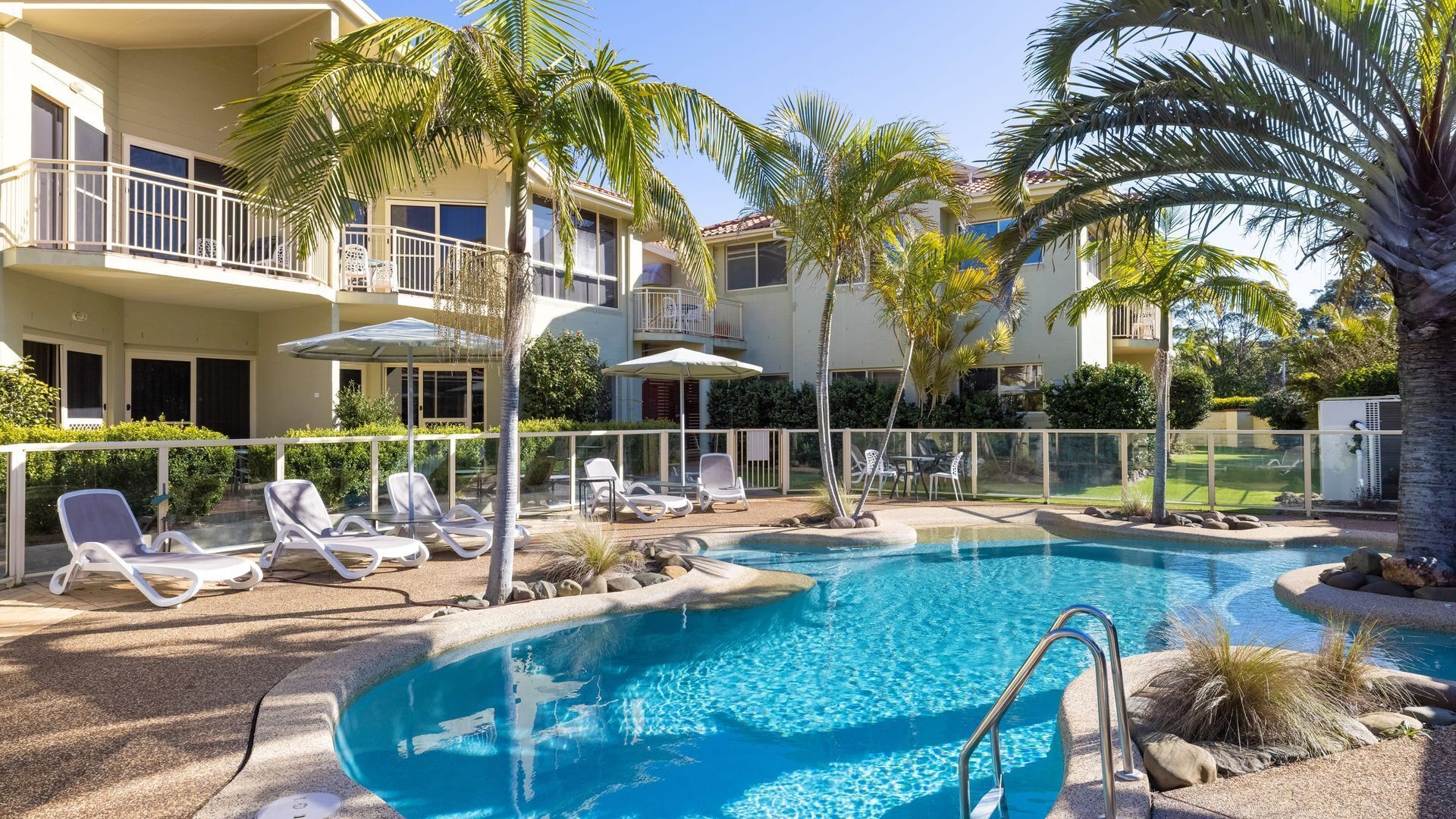A pool area with lounge chairs, palm trees, and a building with balconies.