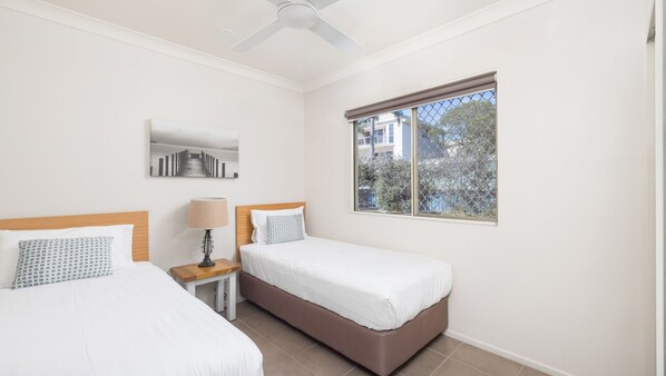 Habitación de hotel con dos camas individuales, una mesita de noche con lámpara, ventilador de techo y ventana con vistas a árboles y edificios.