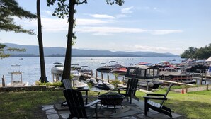 A lakeside patio with Adirondack chairs, a grill, and a dock with boats. - Direct Waterfront Rental  (Lake George)