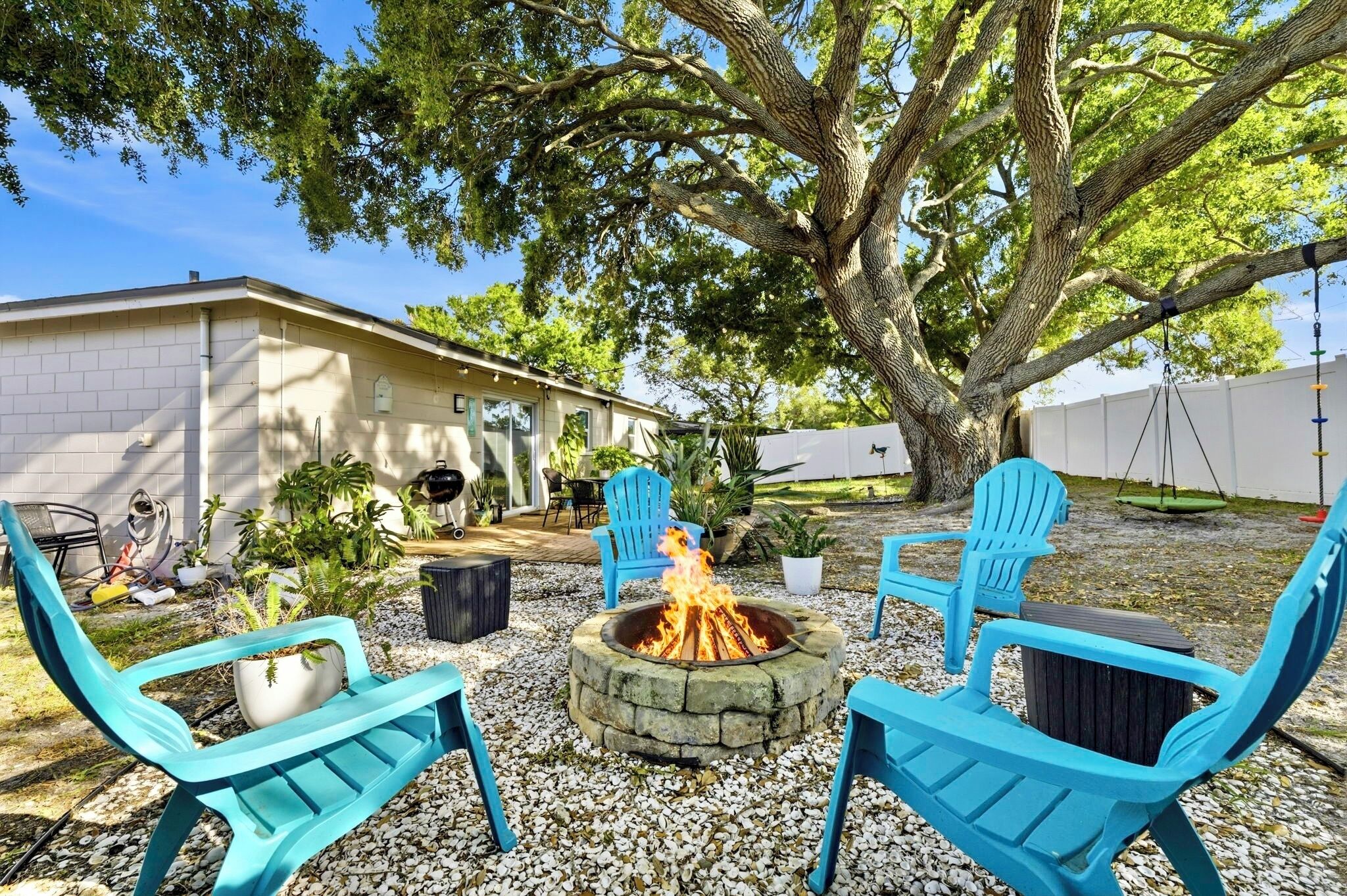 A backyard with a stone fire pit, blue chairs, and a large tree.