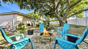A backyard with a stone fire pit, blue chairs, and a large tree.