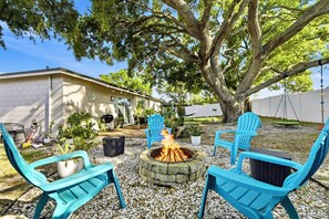 A backyard with a stone fire pit, blue chairs, and a large tree.