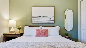A neatly made bed with white linens, a pink pillow, a framed picture of a surfer on the beach, a bedside lamp, and a small potted plant.