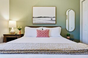 A neatly made bed with white linens, a pink pillow, a framed picture of a surfer on the beach, a bedside lamp, and a small potted plant.