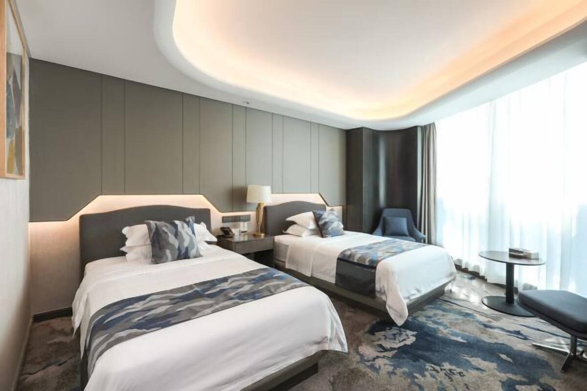 Een hotelkamer met twee bedden, een klein tafeltje, een stoel en een groot raam met gordijnen.