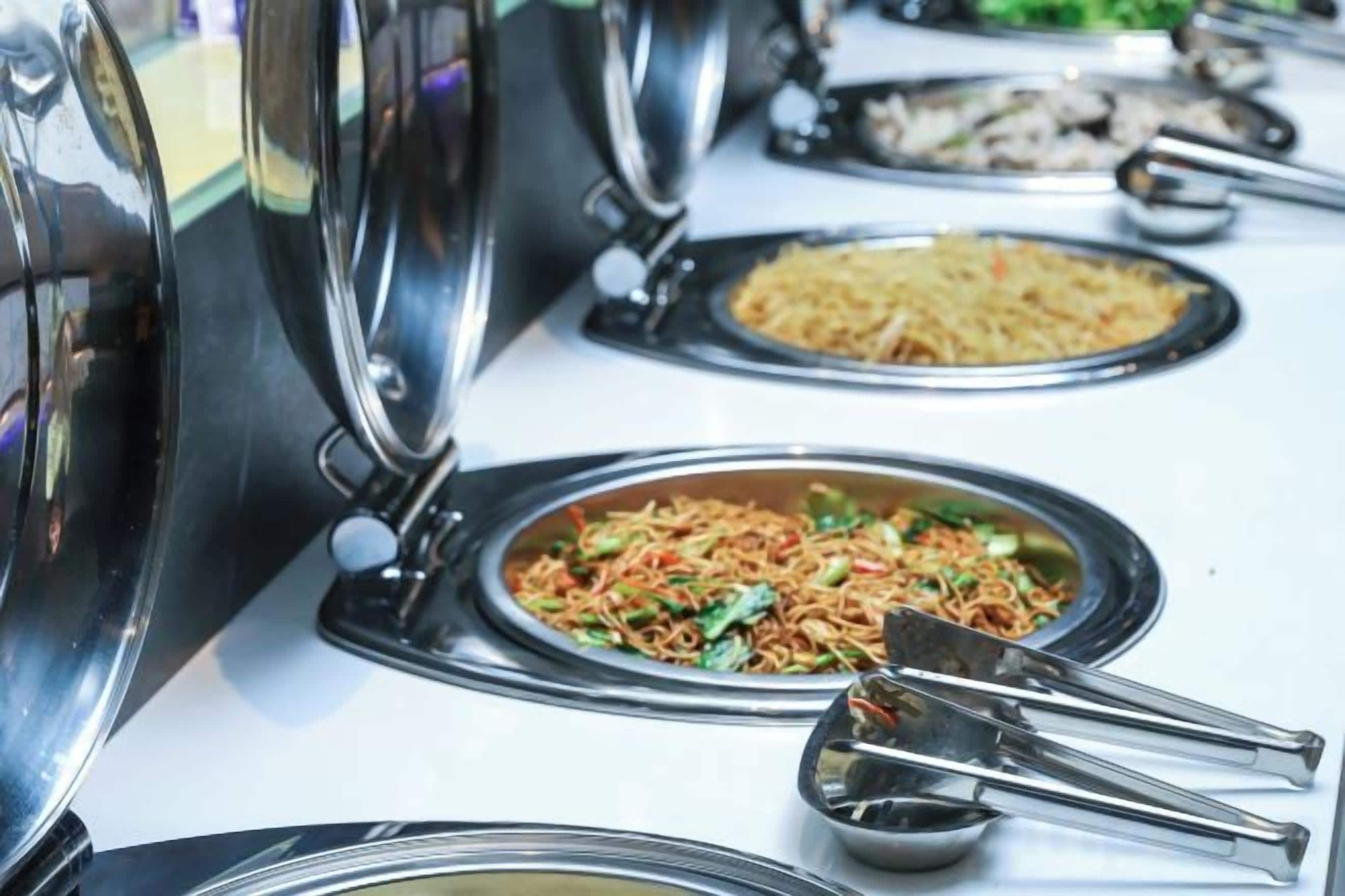 Una mesa de buffet con diversos platos, entre ellos fideos y arroz frito, y utensilios para servir.
