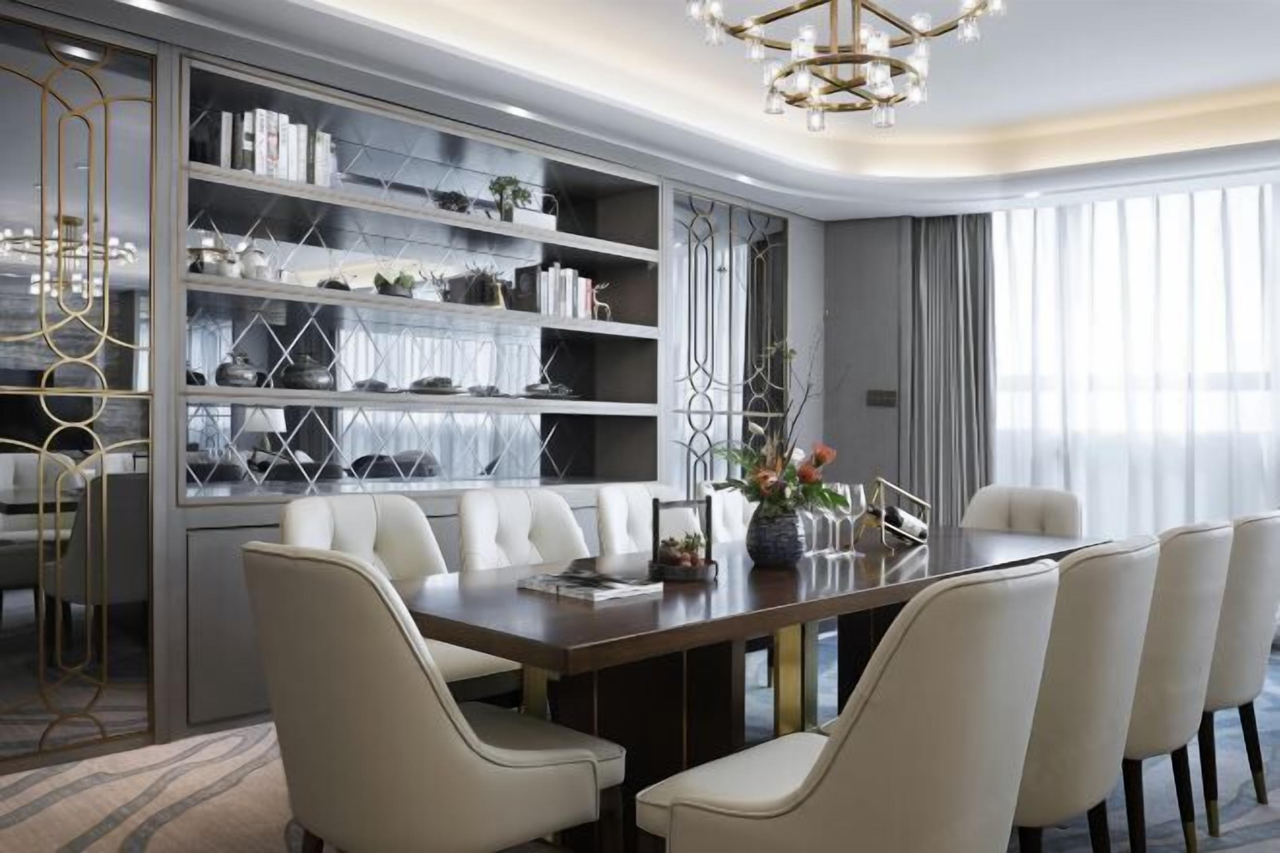A modern dining area with a long table, white upholstered chairs, and a built-in bookshelf with decorative items.