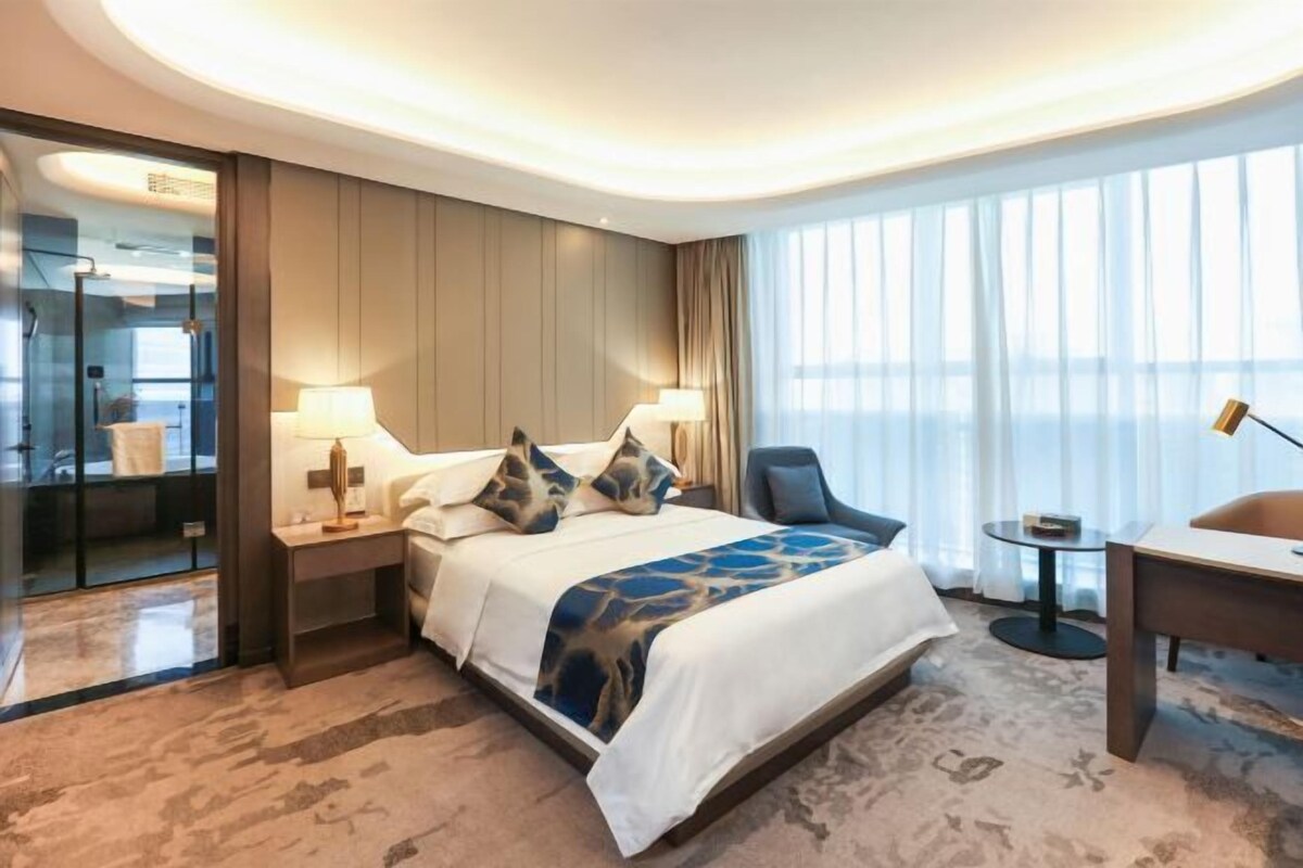 Ein Hotelzimmer mit Bett, Nachttischen, einem Schreibtisch, einem Sessel und einem großen Fenster mit Vorhängen.