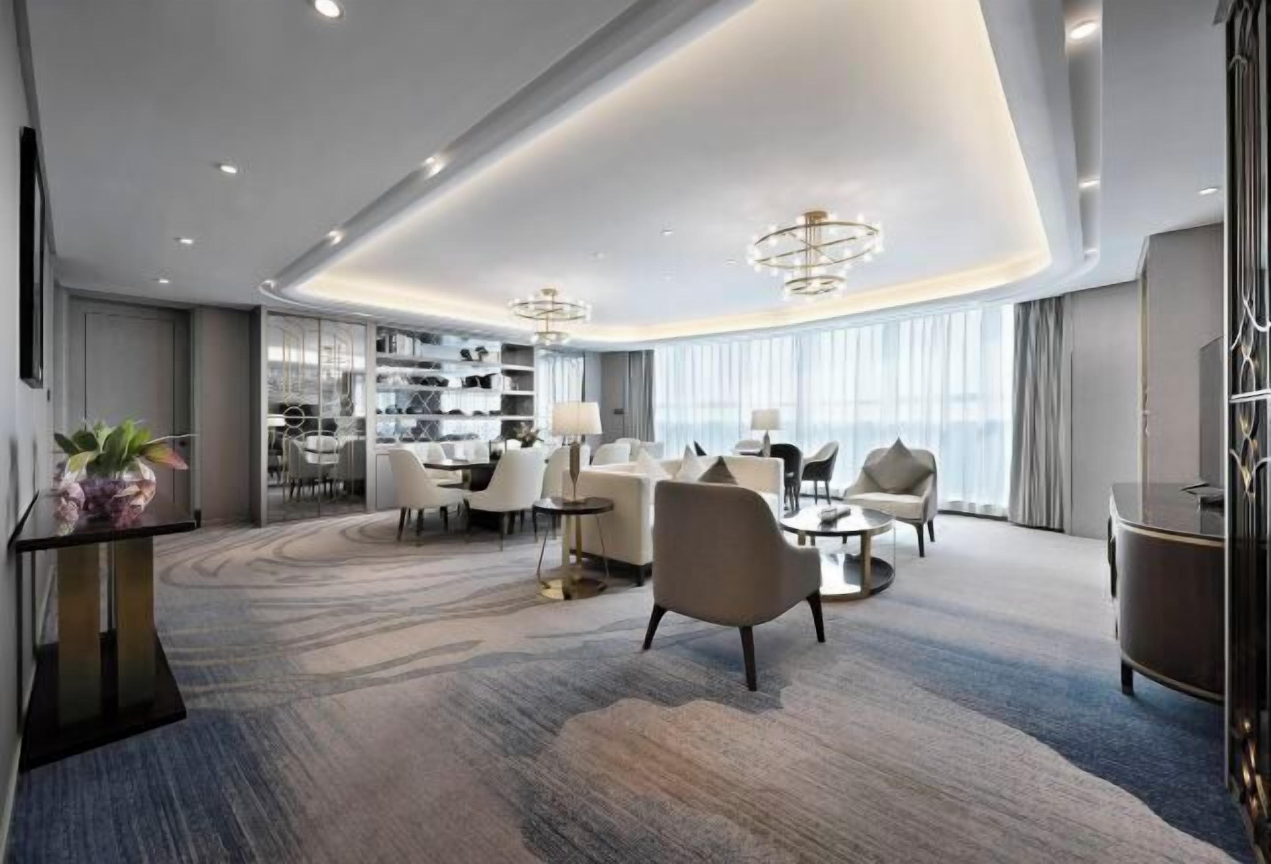 A modern hotel lobby with a curved ceiling, a seating area with sofas and armchairs, a coffee table, and a large window with curtains.