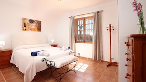 Um quarto com cama, mesas de cabeceira, cômoda de madeira, janela com cortinas e um quadro na parede.