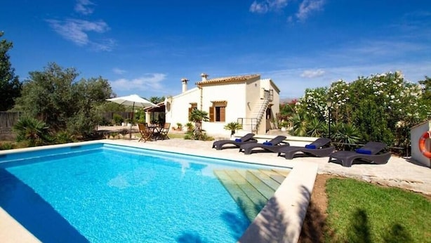 Piscina com espreguiçadeiras, um edifício branco com telhado em azulejos e vegetação ao redor.