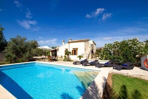 A swimming pool with lounge chairs, a white building with a tiled roof, and surrounding greenery.