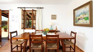 Uma sala de jantar com uma mesa e cadeiras de madeira, uma janela com cortinas, um quadro na parede e uma pequena mesa com abajur e objetos decorativos.