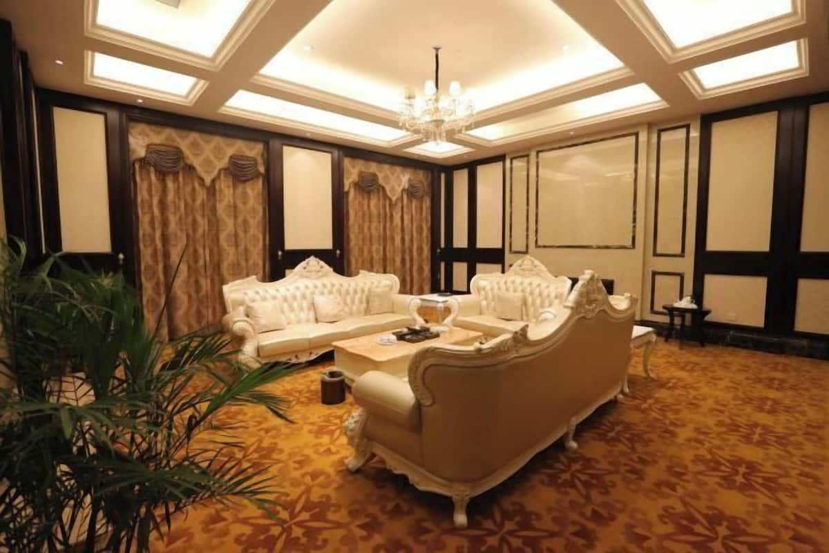 A luxurious living room with a white sofa set, a coffee table, and a chandelier. The room has a patterned carpet and large windows with curtains.
