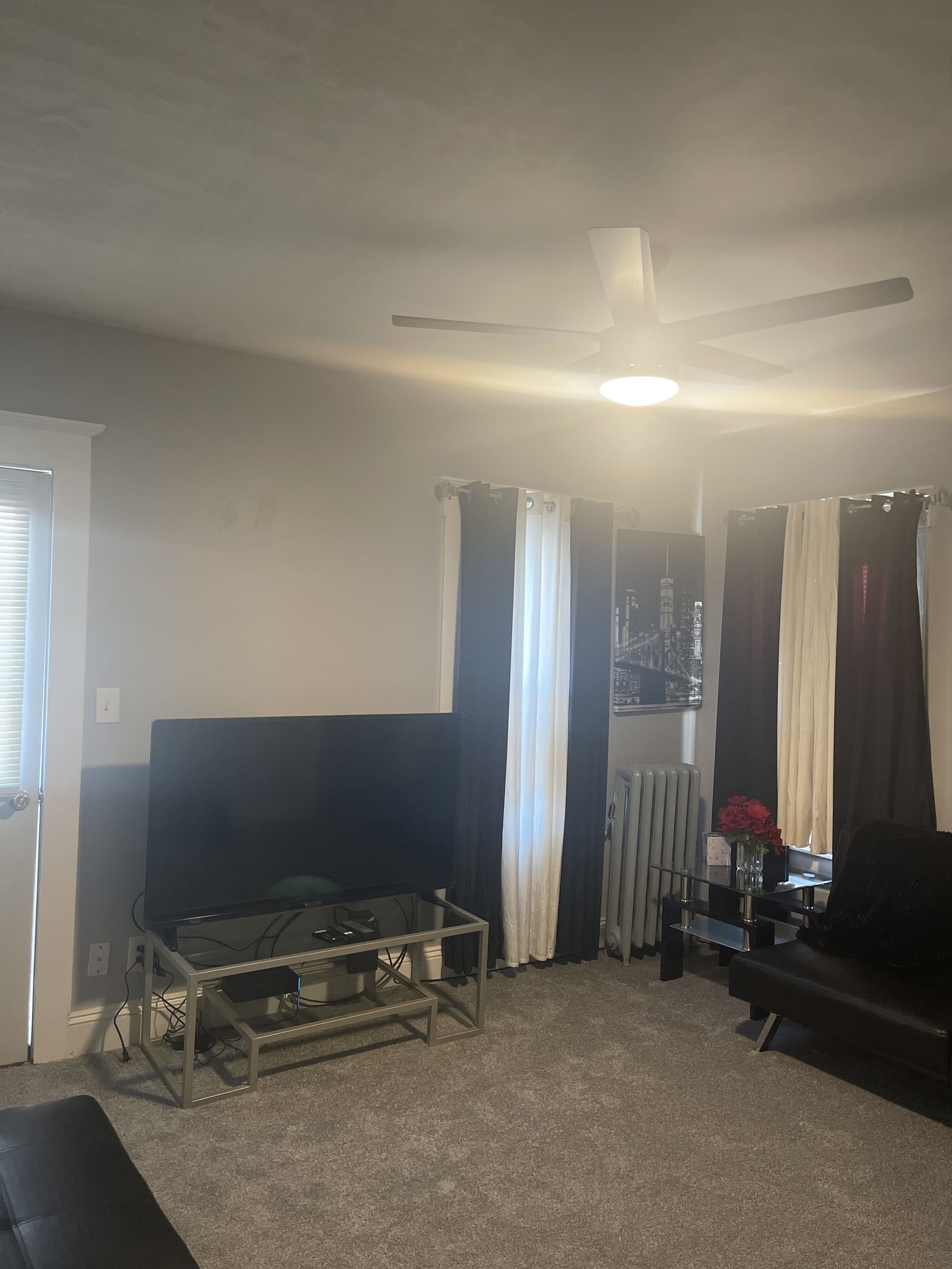 A living room with a flat-screen TV on a glass table, a ceiling fan, and a radiator.