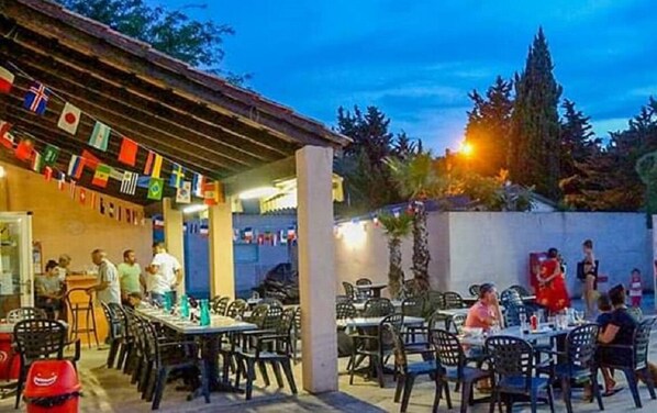 An outdoor dining area with tables and chairs, flags hanging above, and a building with a tiled roof in the background.