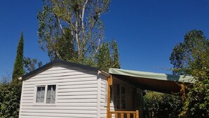 A small, single-story cabin with a porch and a covered area, surrounded by trees. - Mobile home in camping Le mas de l'isle (LUNEL)