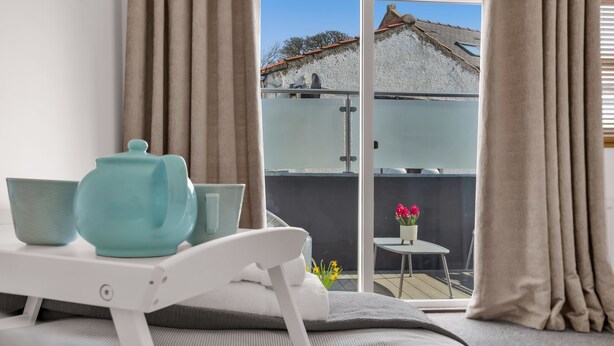 A bedroom with a bed, a bedside table with a kettle and bowls, a view of a balcony with a table and flowers, and curtains. - Shorefront Anstruther Home - 2 Bedrooms (Anstruther)