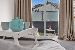 A bedroom with a bed, a bedside table with a kettle and bowls, a view of a balcony with a table and flowers, and curtains. - Shorefront Anstruther Home - 2 Bedrooms (Anstruther)