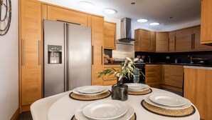 A modern kitchen with wooden cabinets, a stainless steel refrigerator, and a dining table set with plates and a vase of flowers. - Shorefront Anstruther Home - 2 Bedrooms (Anstruther)
