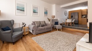 A modern living room with a grey sofa, a grey armchair, and a small table with a lamp. There is a kitchen area with wooden cabinets and a refrigerator. - Shorefront Anstruther Home - 2 Bedrooms (Anstruther)