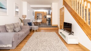 A modern living room with a grey sofa, a coffee table, and a TV on a stand. There is a staircase with wooden railings leading to an upper floor. - Shorefront Anstruther Home - 2 Bedrooms (Anstruther)
