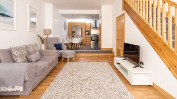 Una sala de estar moderna con un sofá gris, una mesa de centro y un televisor sobre un mueble bajo. Se ve una escalera con barandilla de madera que sube a un nivel superior.