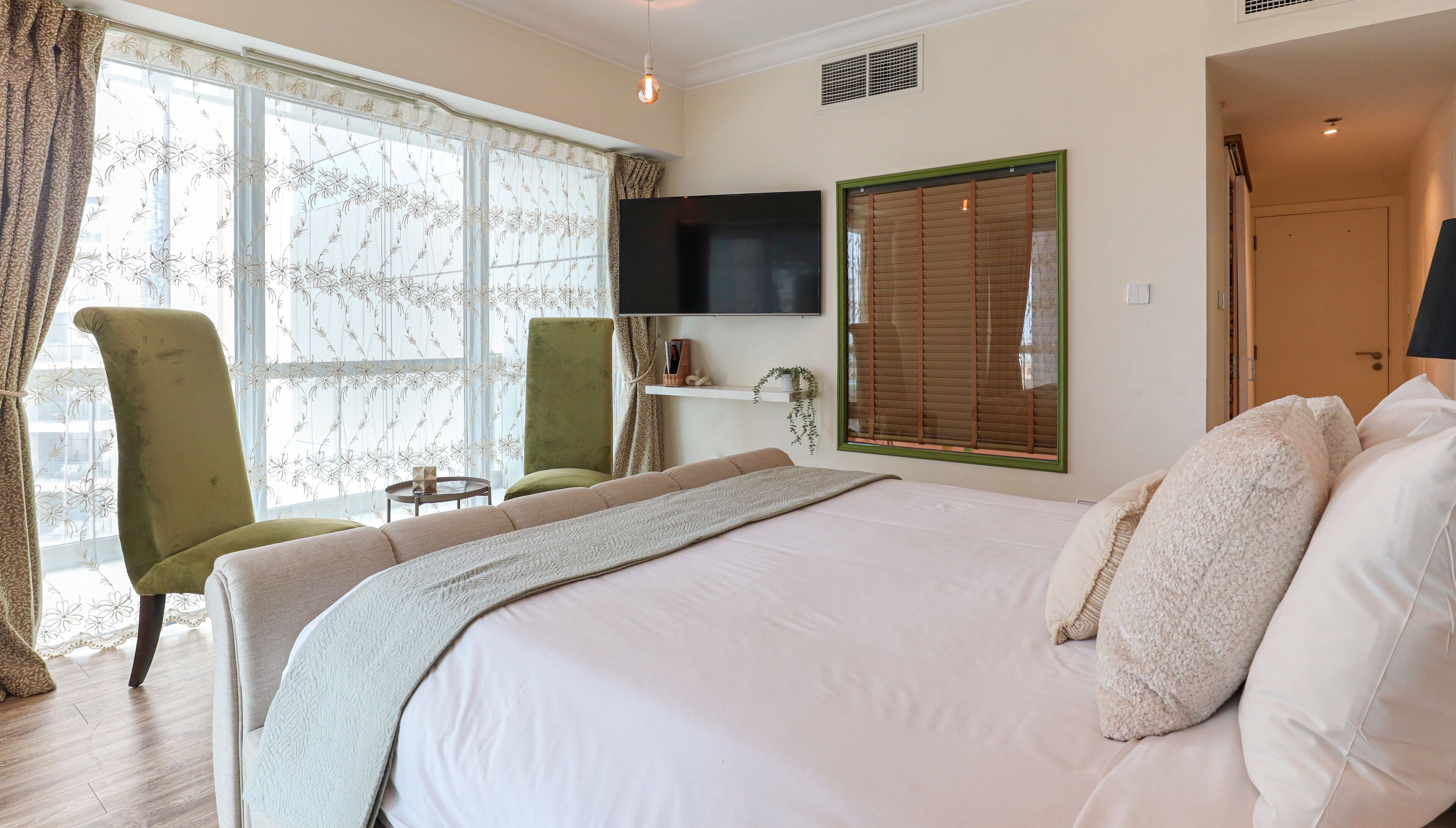 A bedroom with a large bed, a television mounted on the wall, a chair, a small table, and a window with curtains.