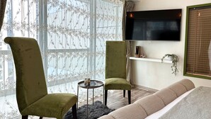 Un salon avec deux fauteuils verts, un canapé beige, une petite table ronde sur laquelle se trouve un magazine, et un téléviseur fixé au mur.