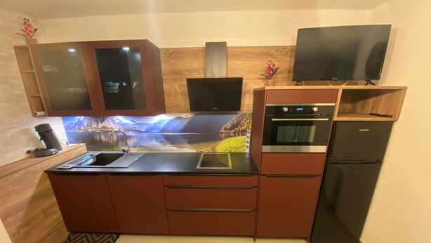 A compact kitchen with a built-in stove, oven, and refrigerator, a sink, and a television mounted above the counter.