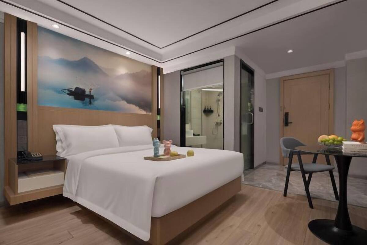Una habitación de hotel moderna con cama, un cuadro de un helicóptero, una mesita con frutas y un baño visible a través de una puerta de vidrio.