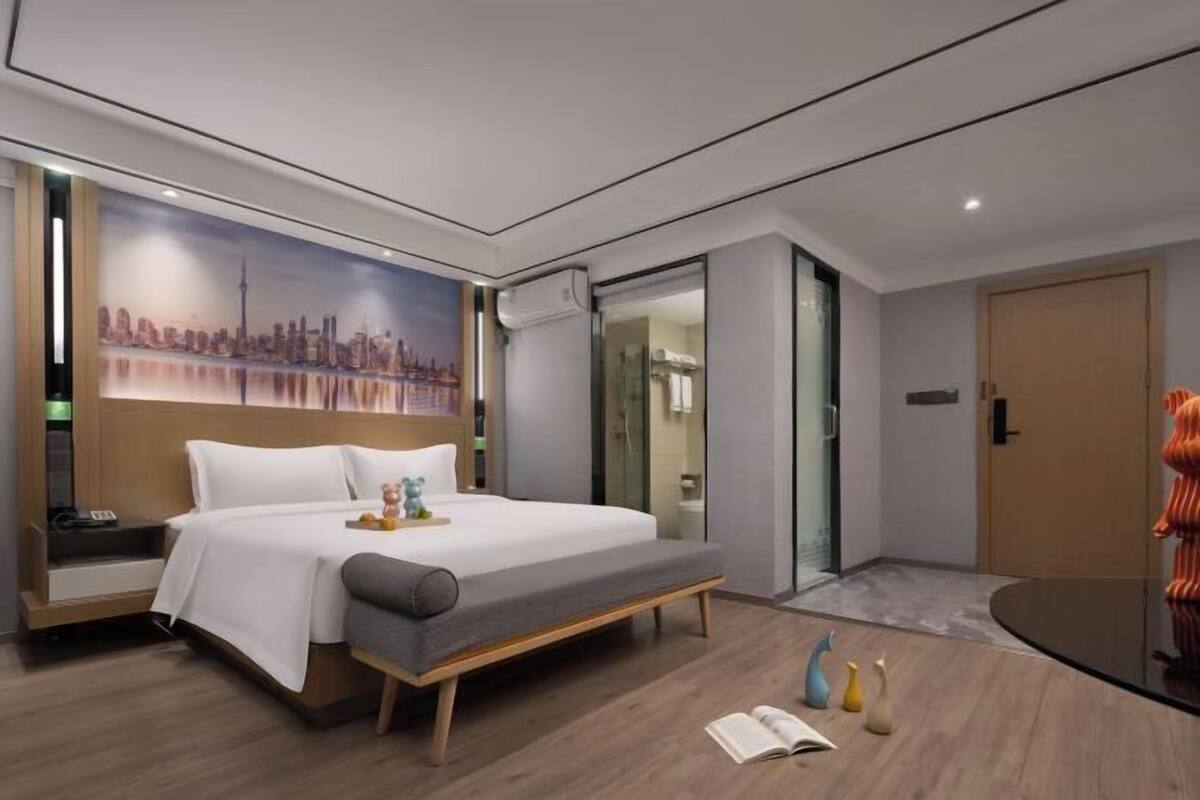 Una habitación de hotel moderna con una cama, un mural con vista a la ciudad, zona de ducha con mampara de vidrio y un pequeño rincón de lectura con libros y objetos decorativos.