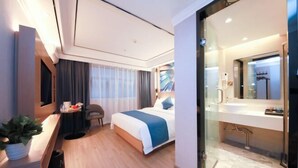 Une chambre d’hôtel moderne comprenant un lit, une table de chevet, une plante en pot et une salle de bain fermée par des parois de verre, avec un lavabo et un miroir.